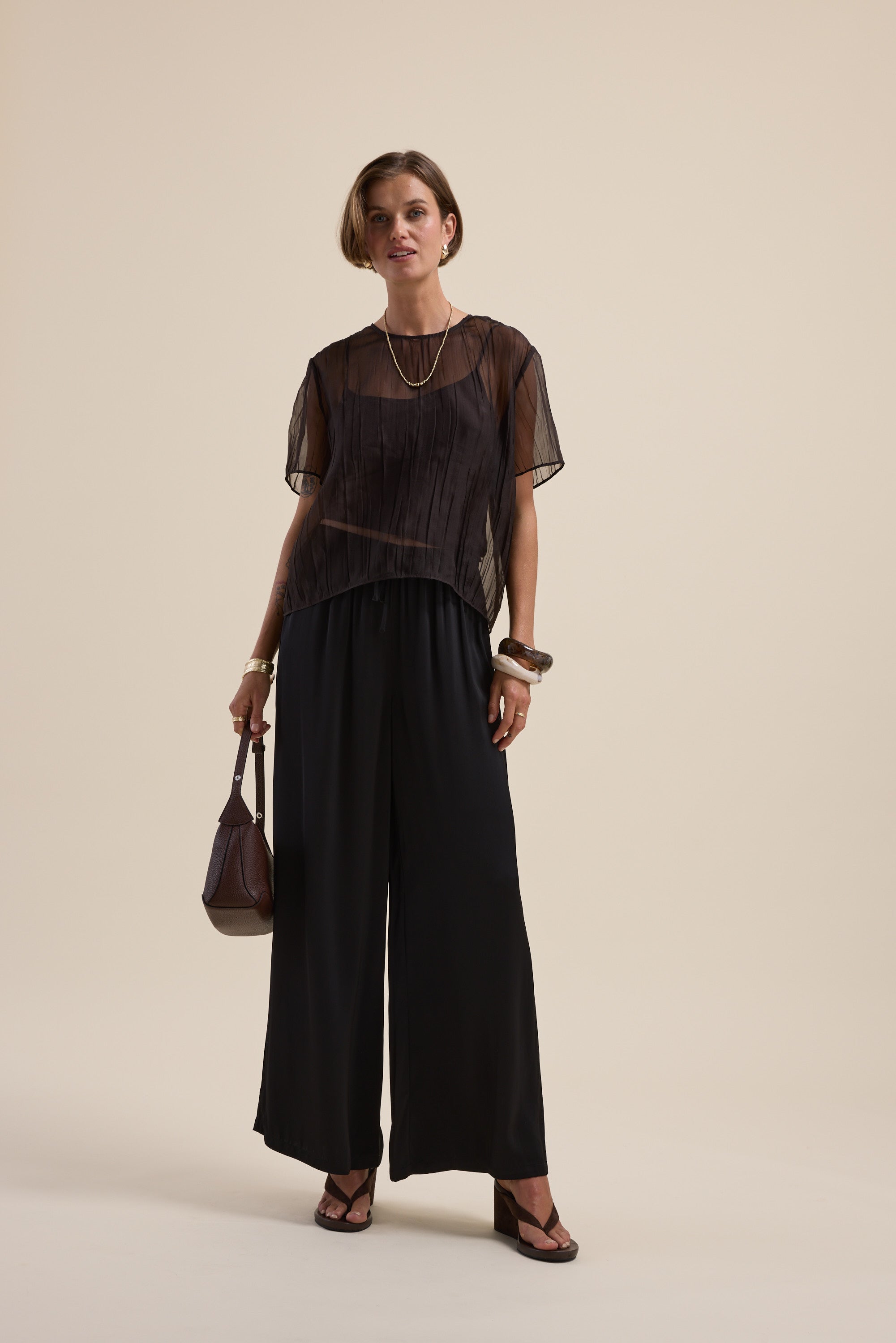 Woman in Max's Cass Sheer Top with detachable cami, wide pants, brown bag, and sandals.