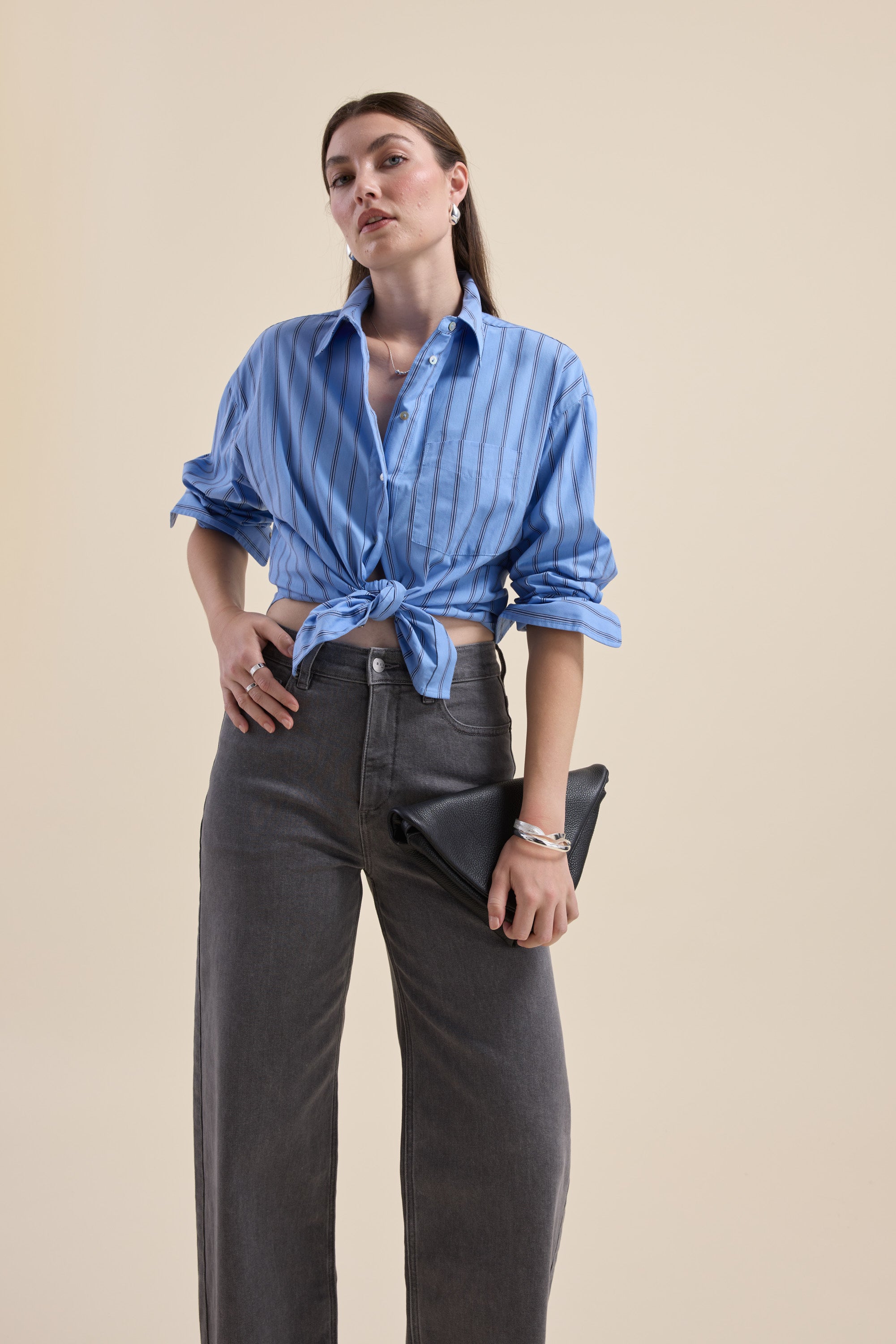 Woman wears Max Cameron Oversized Shirt and gray pants, holding a black clutch against beige.