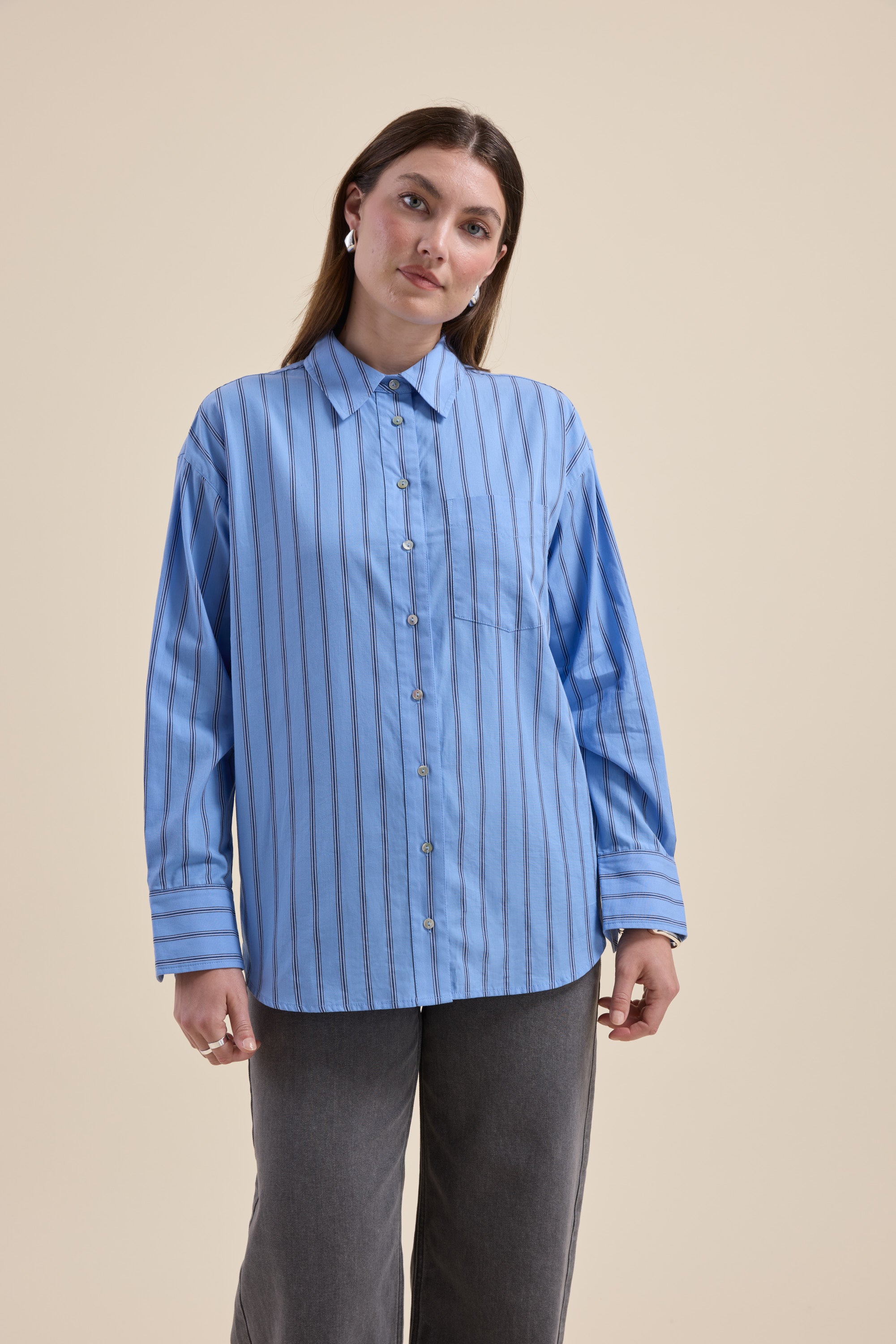 Woman wears Max Cameron Oversized Shirt, blue striped, with gray pants against beige background.
