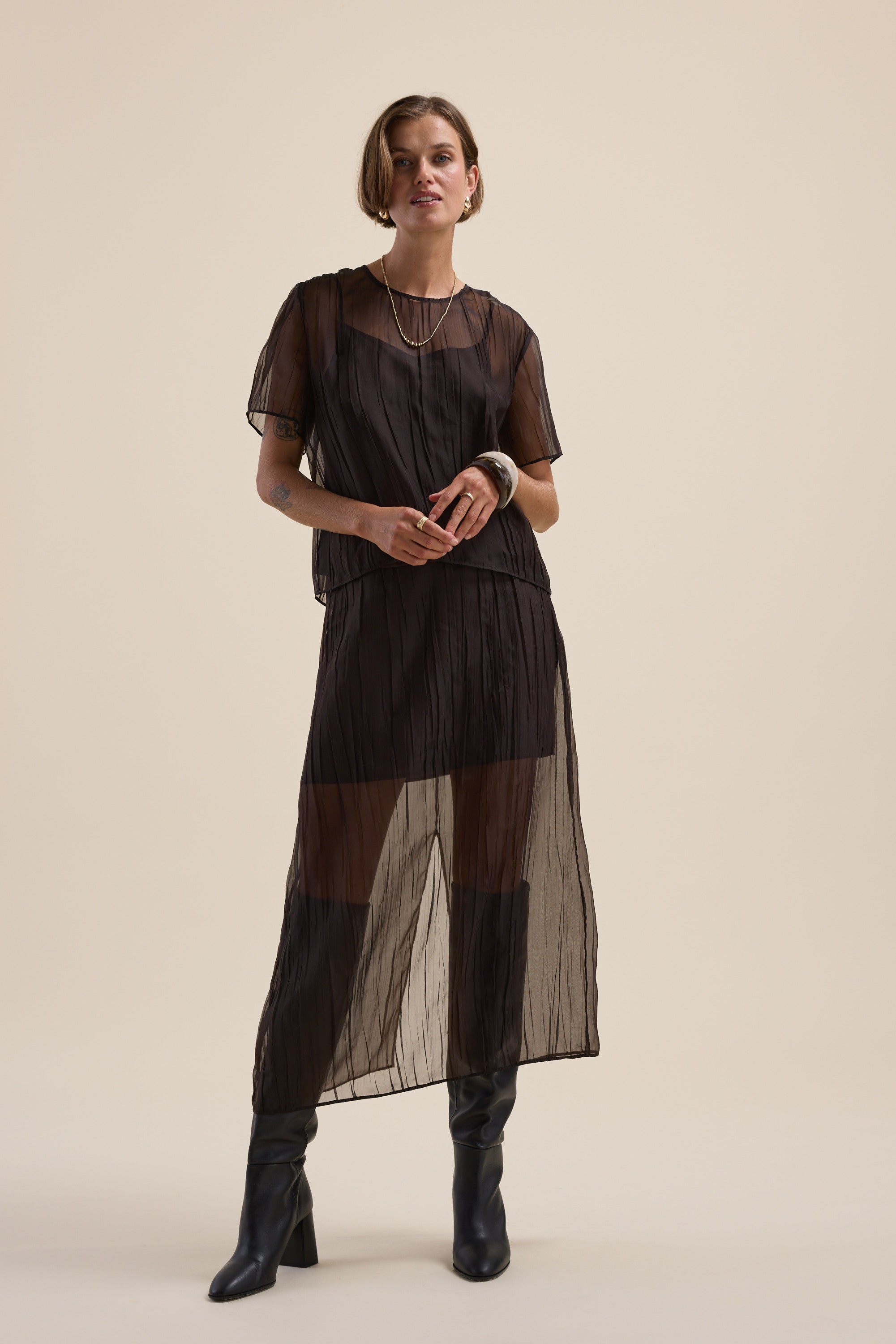 Model in Cass Sheer Top by Max, black boots, short hair, set against a beige background.