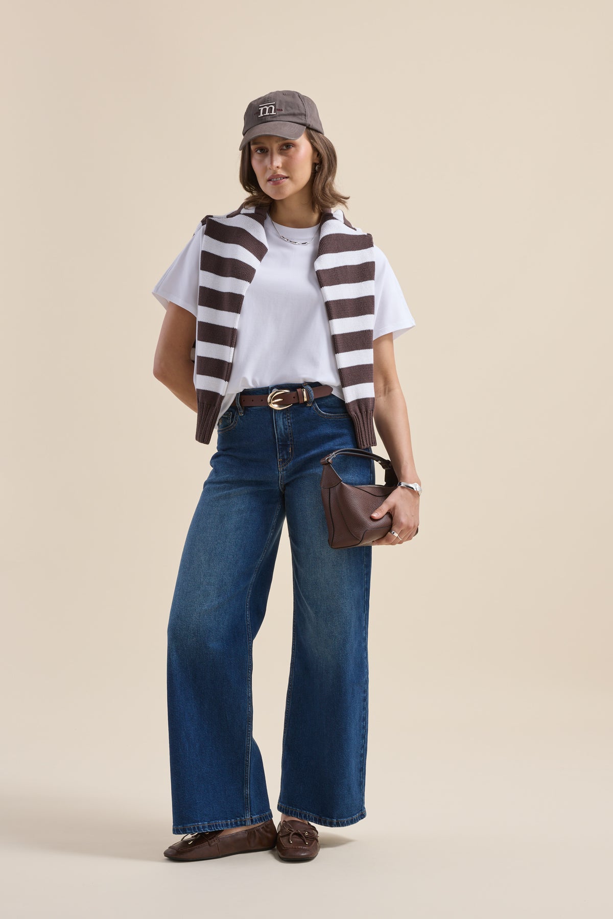 Woman in wide-leg jeans, Elementary tee, striped sweater, cap, and holding a purse.