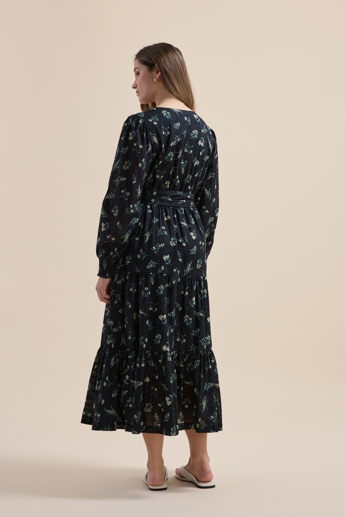Woman in Elementary Tiered Dress with daisy sprig print and sandals, facing away, plain backdrop.