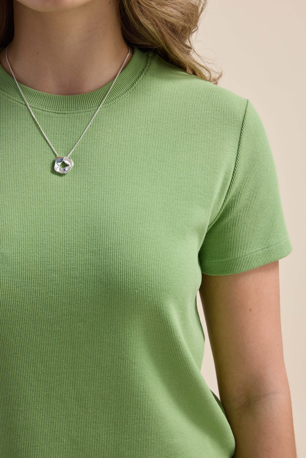 Person wears Elementary Ribbed Boxy Tee in green, silver pendant necklace, beige background.