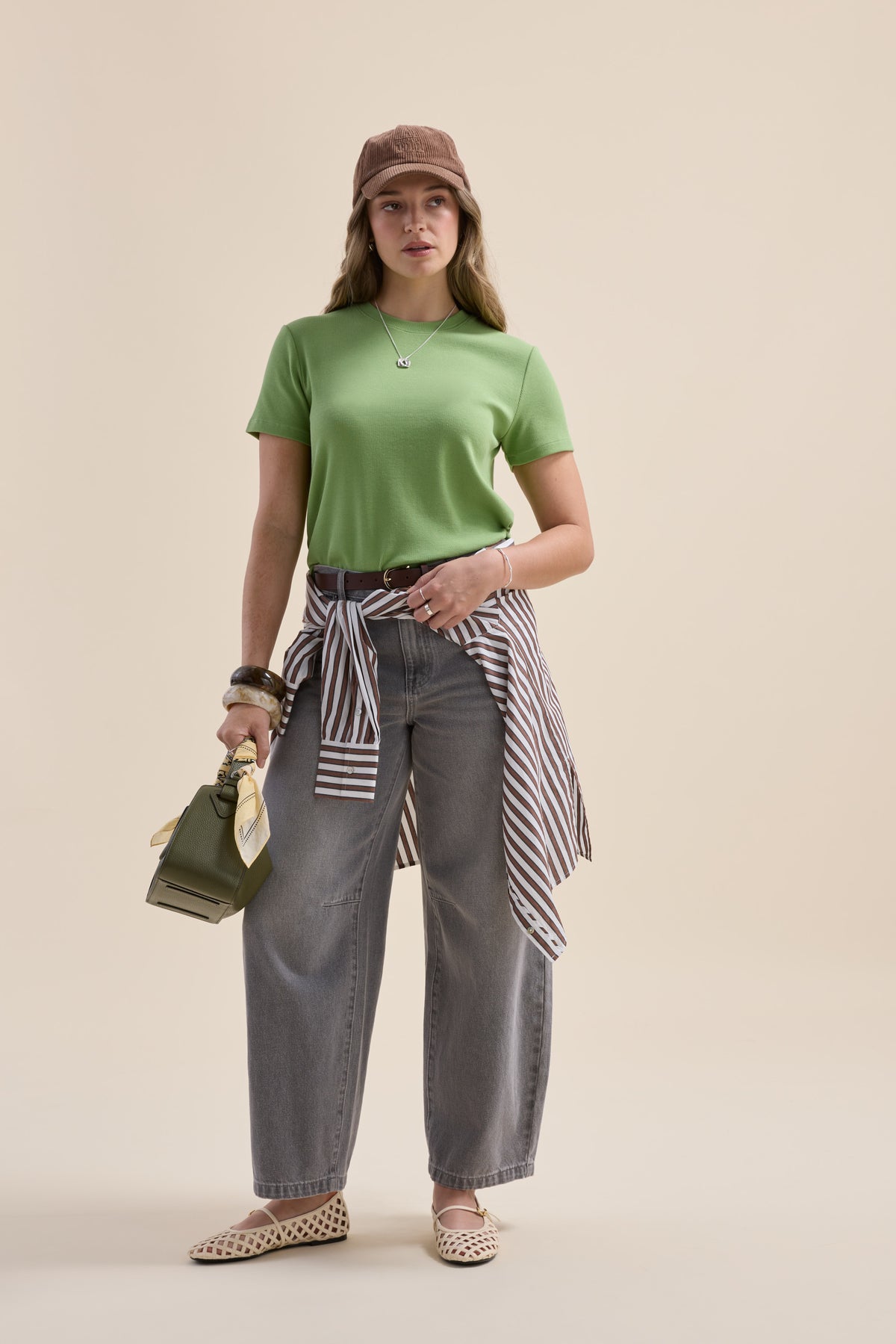 Woman in green Elementary Ribbed Boxy Tee, gray jeans, brown cap, bag, striped shirt at waist.