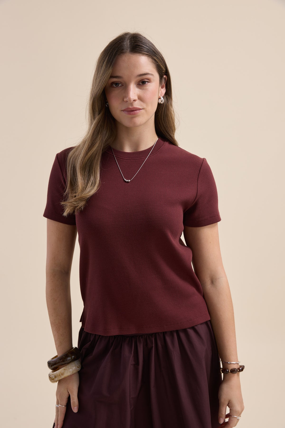 A woman with long brown hair wears the Elementary Ribbed Boxy Tee in maroon and silver jewelry.