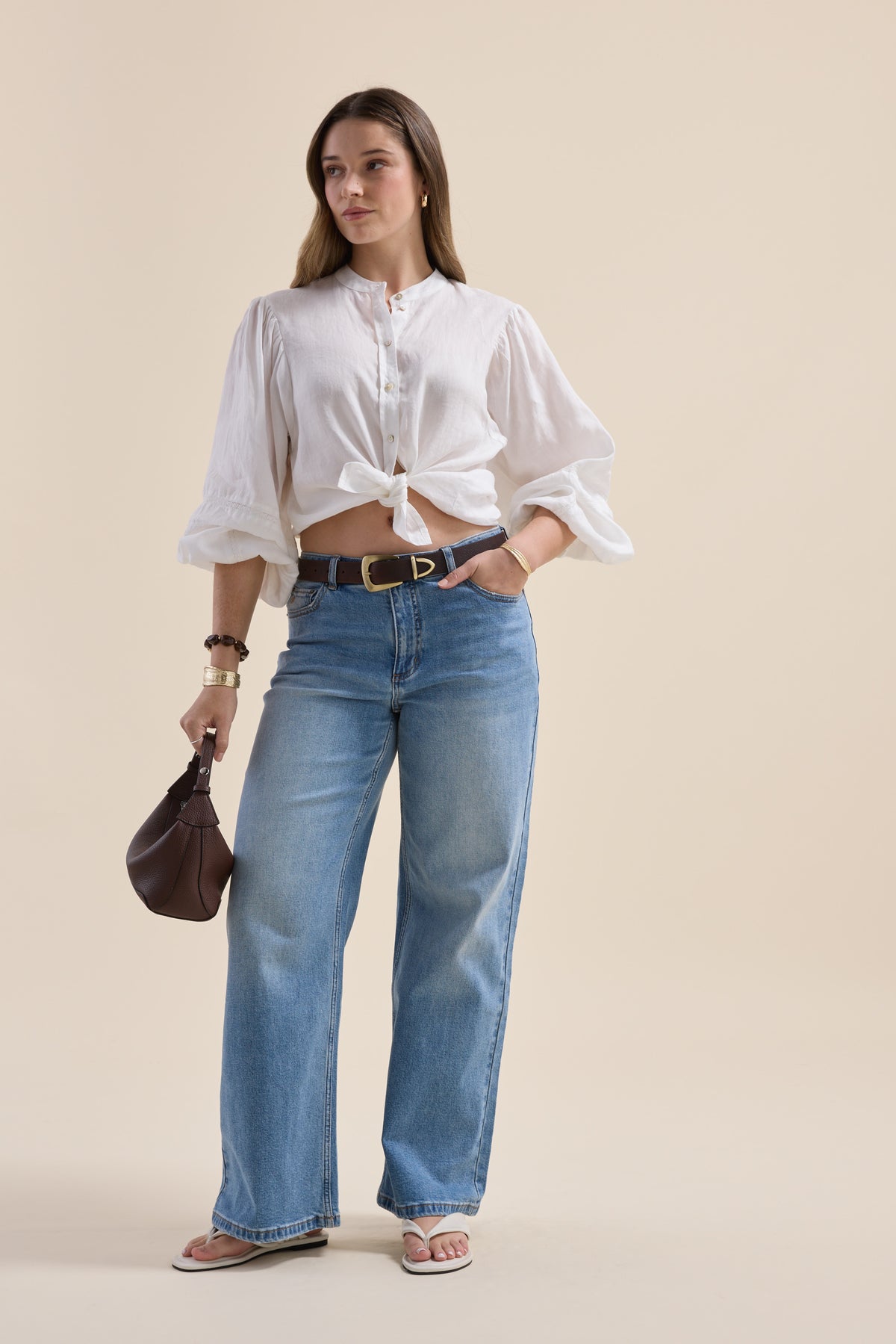 Woman in Max’s Millie Trim Detail Blouse, blue jeans, sandals, with brown bag on beige background.
