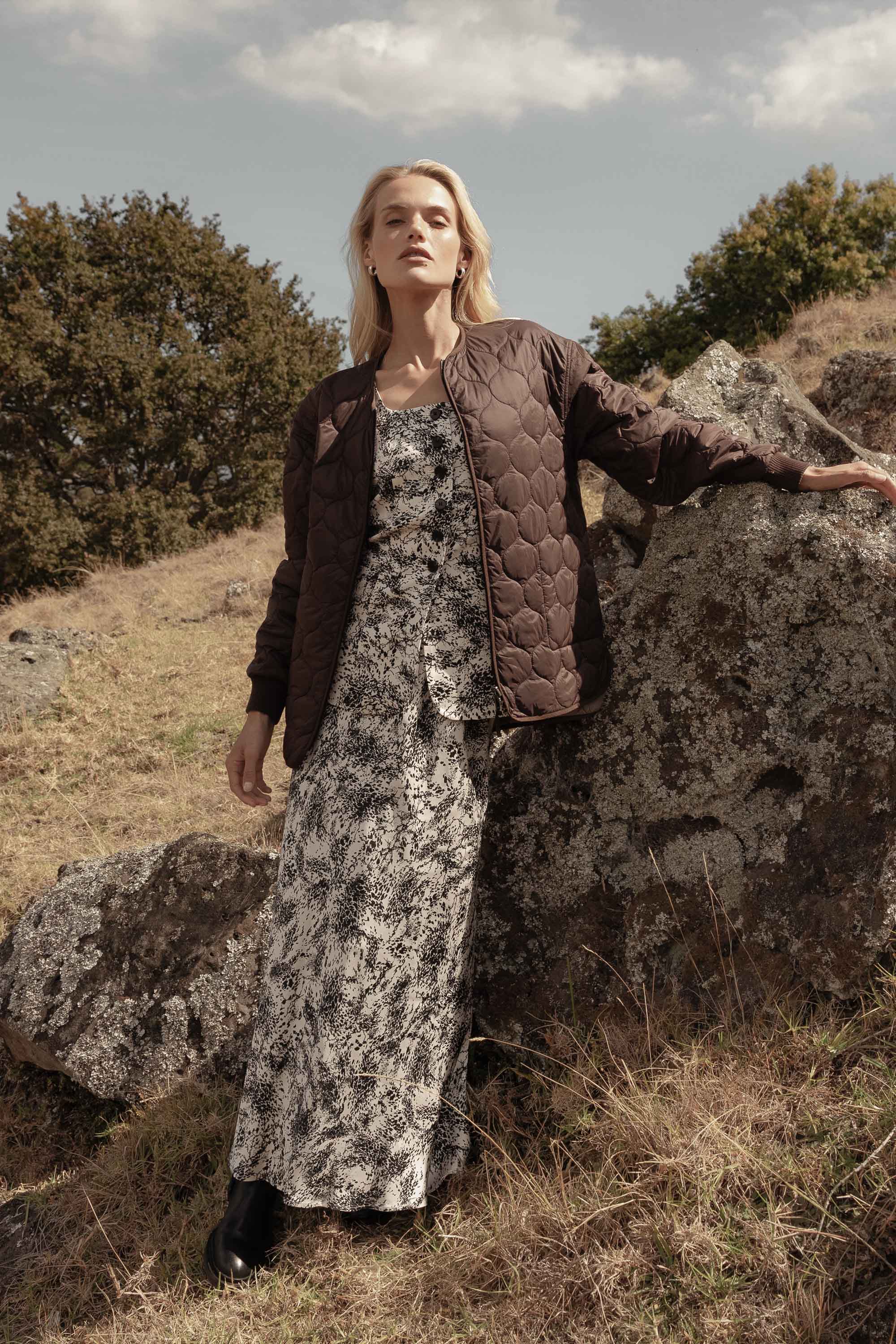 Woman in the Henry Maxi Slip Skirt by Max stands by rocks on a grassy hillside.