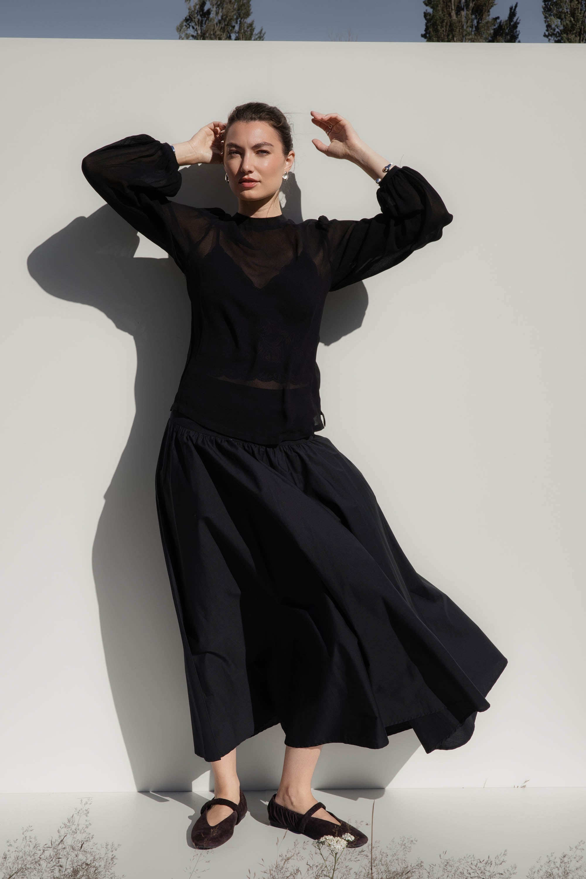 Model wears Max’s The Ultimate Layering Top in black, posing against a white wall in sunlight.