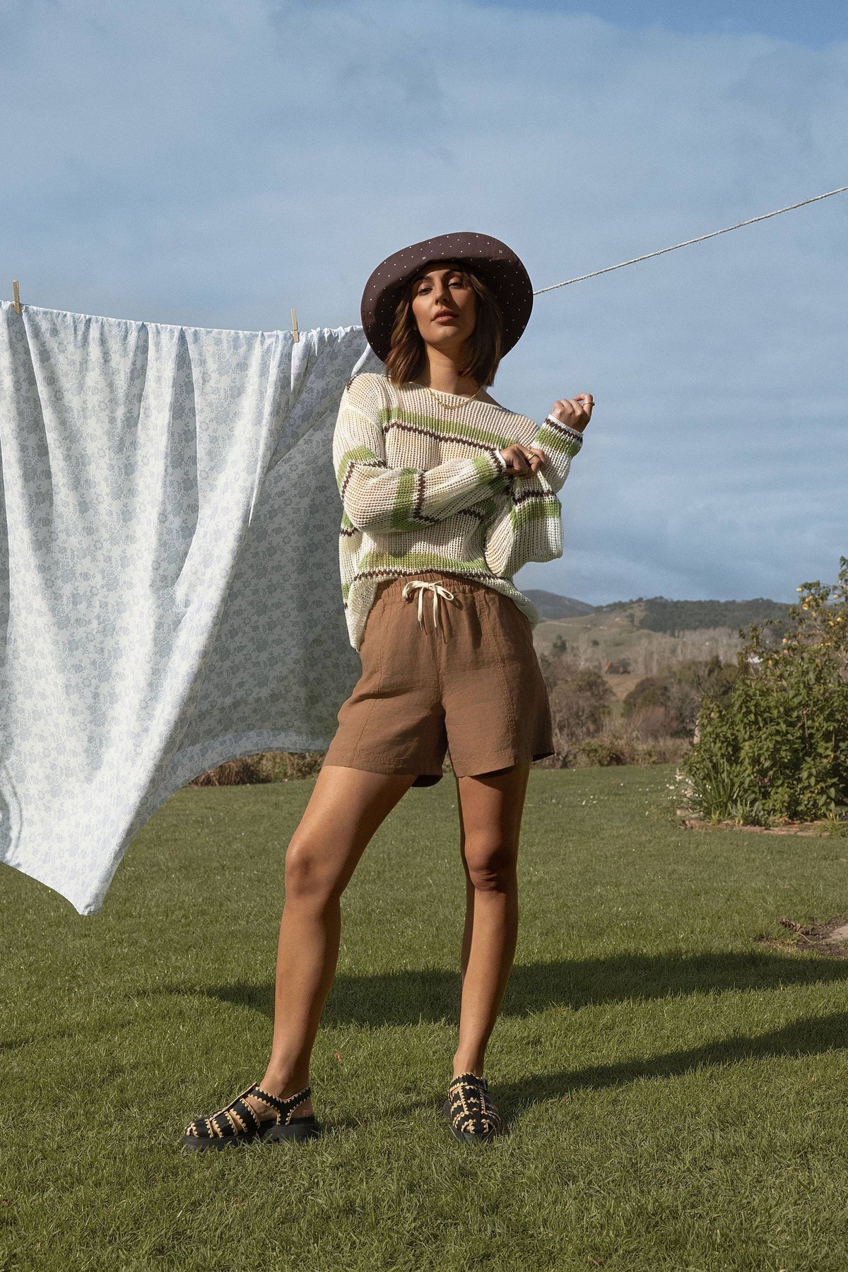 Woman in hat, sweater, and Elementary Linen Short by Elementary stands on grass near laundry line.