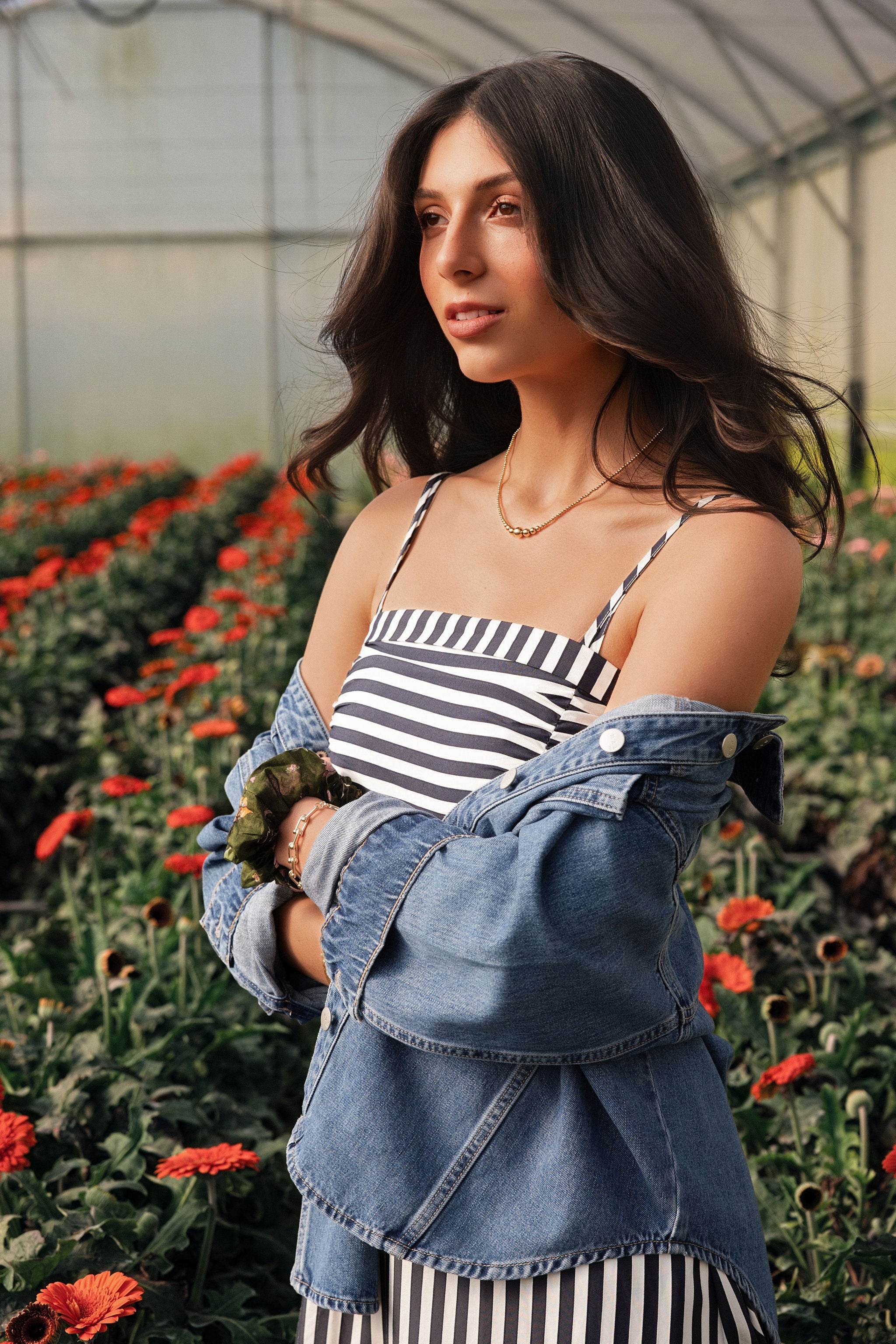 A woman standing in a greenhouse wearing a striped spring dress and a denim jacket.