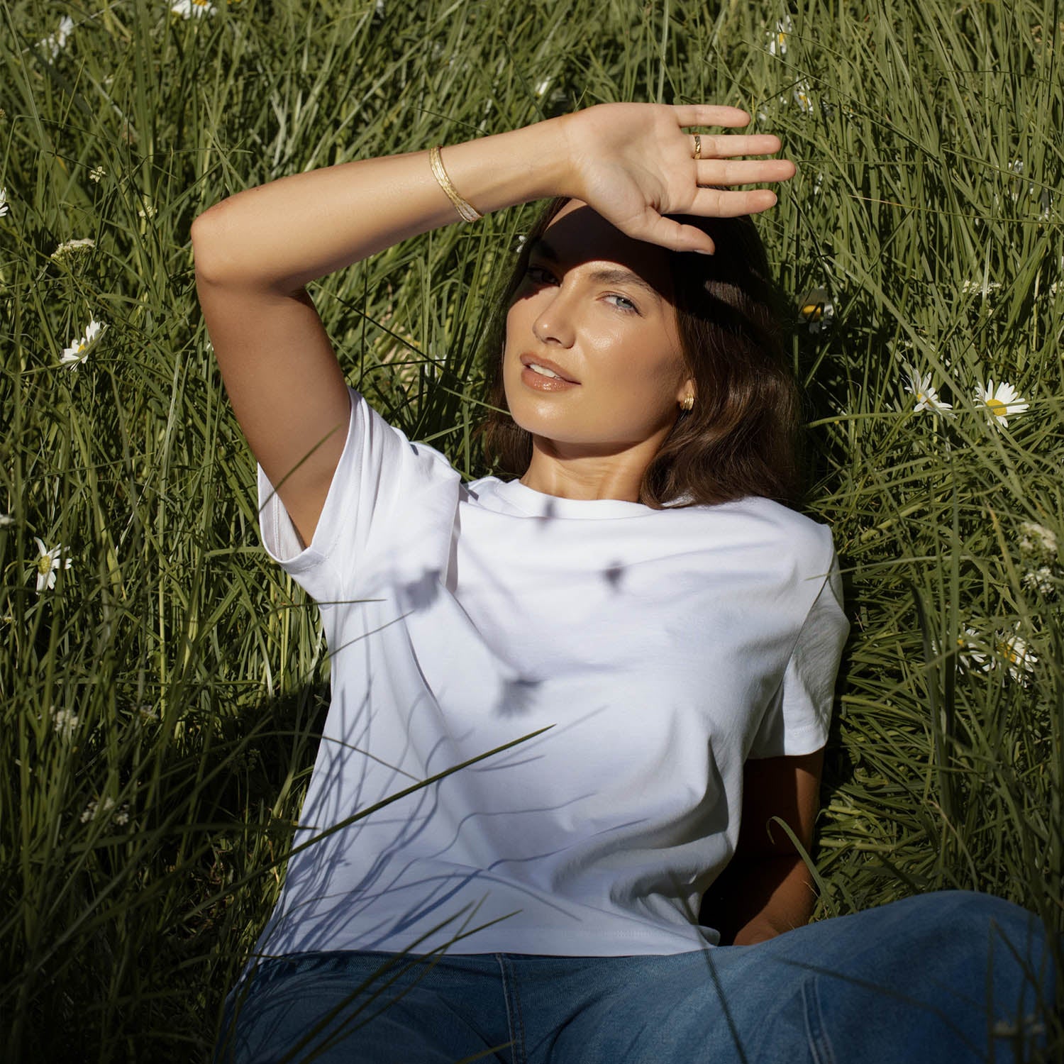 Woman in white shirt lies in NZ grass, shielding her eyes from the sun with her hand.