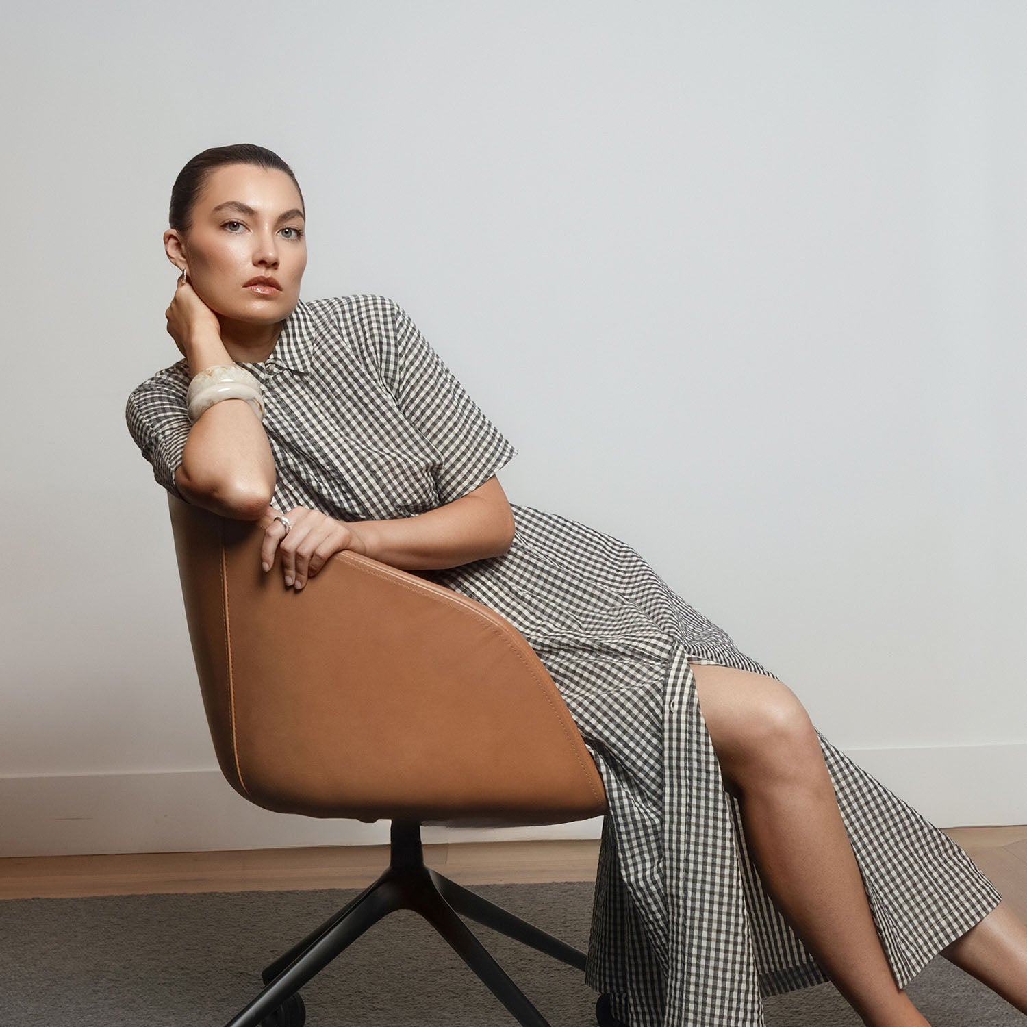 Woman in a checkered dress sits sideways on a tan chair, gazing at the camera in NZ.