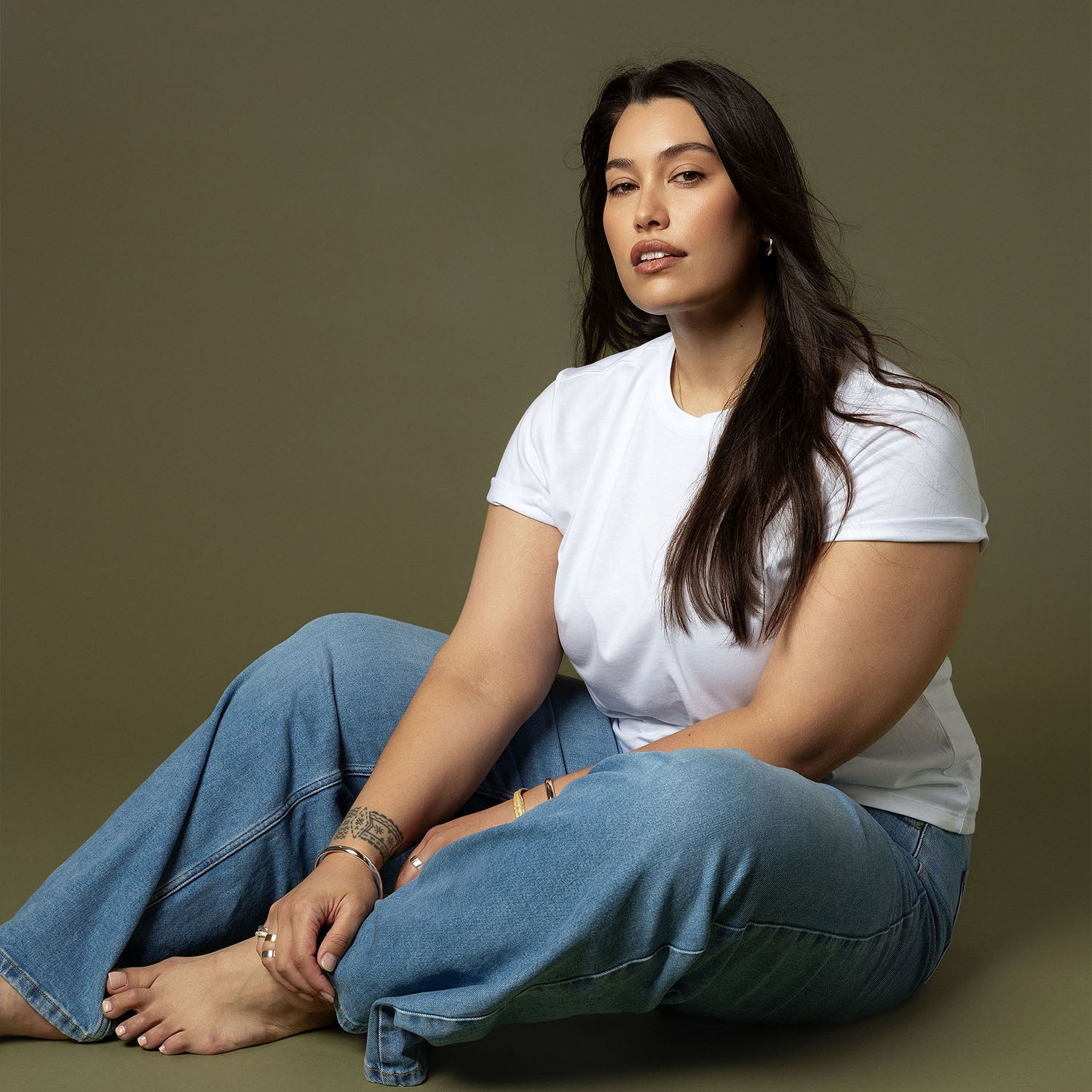 NZ woman in a white t-shirt and jeans sits on the floor against an olive background.