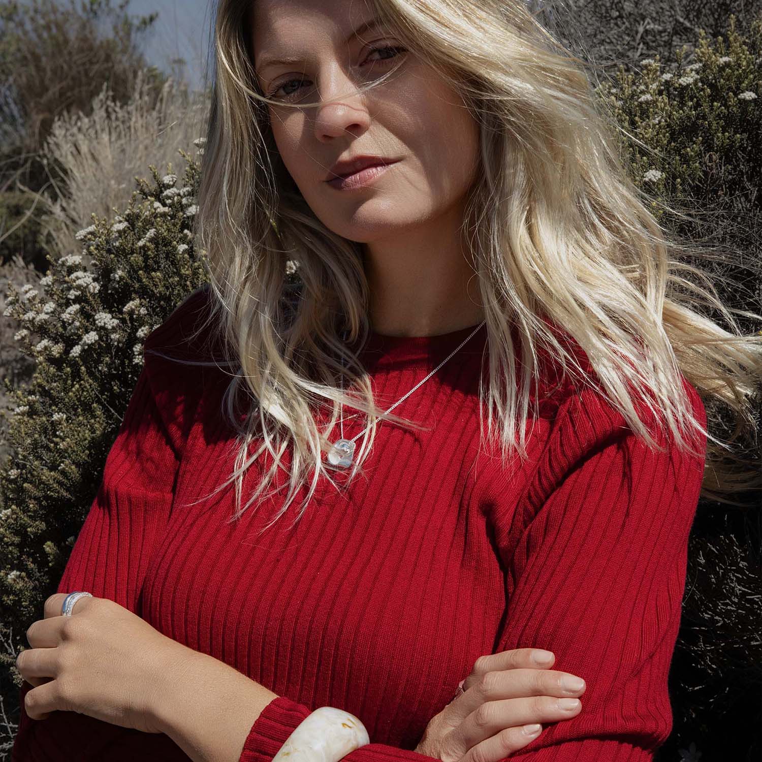 Woman with long blonde hair in a red sweater standing outdoors in NZ with arms crossed.