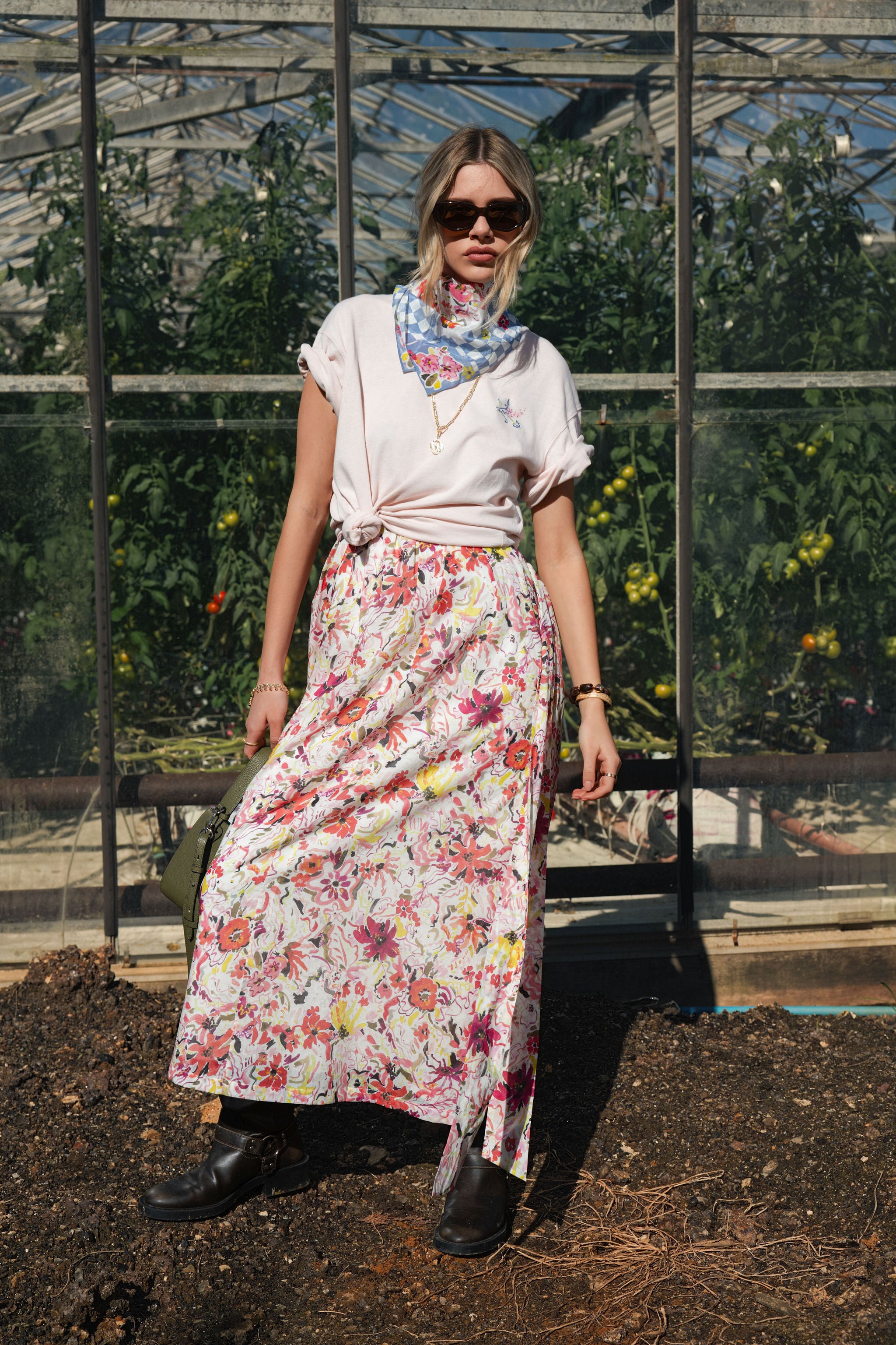 A woman standing in front of a greenhouse wearing a floral spring midi dress layered with an oversized tee and a floral scarf.