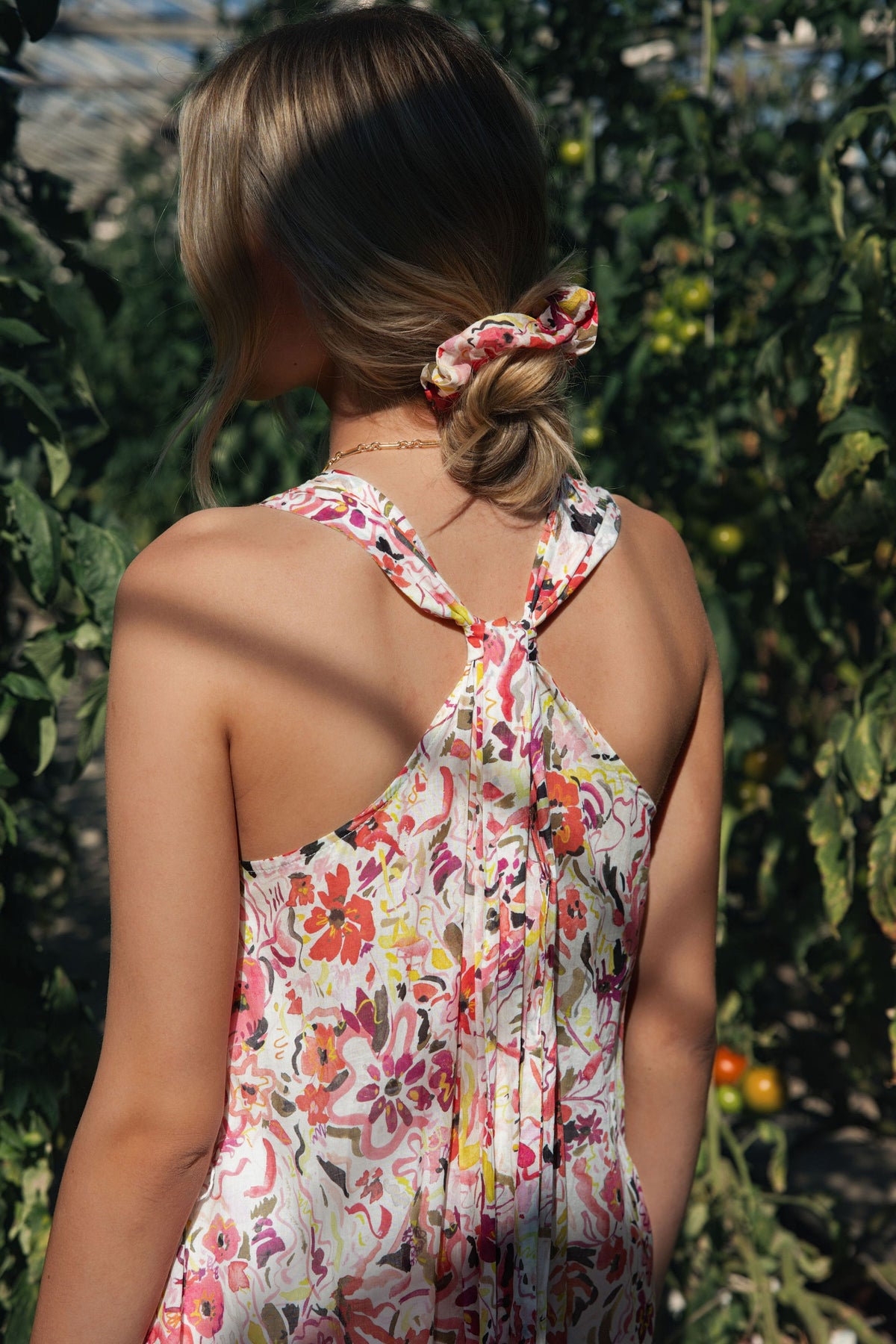 Woman in Max’s Kira Back Detail Dress with matching scrunchie, standing among green plants.
