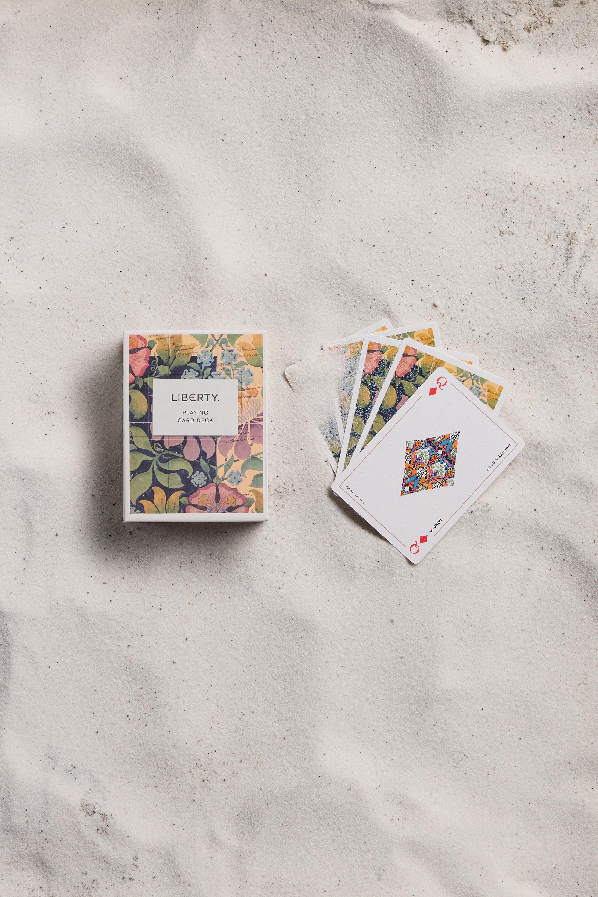 A floral-patterned Liberty Archive Playing Card Deck by Other rests on sand beside fanned-out cards, highlighting their detailed British-inspired artwork.
