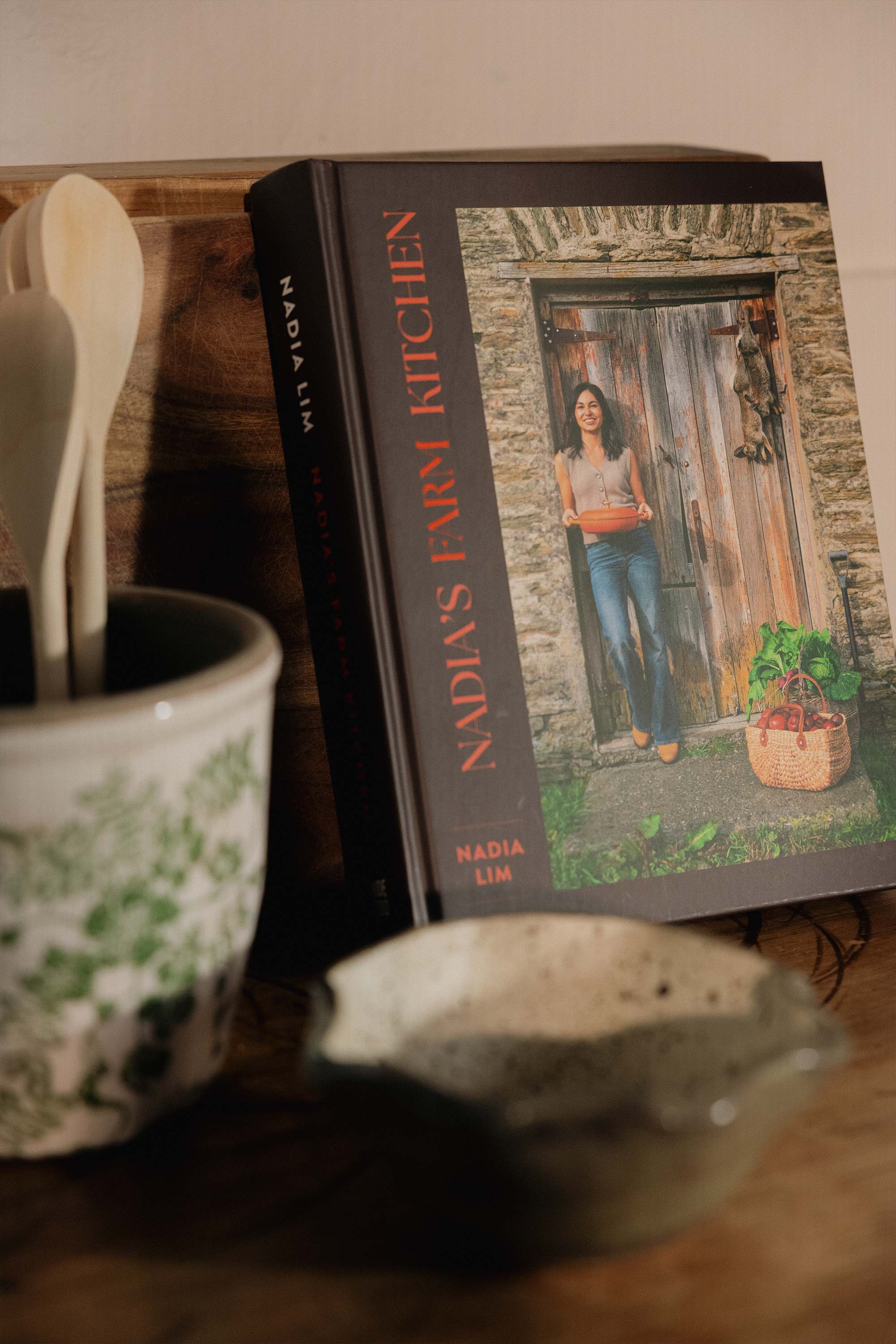 Nadia's Farm Kitchen Book by Other rests against a wooden surface next to a ceramic jar of utensils and a speckled bowl, its cover showing Nadia outside a rustic door—ideal for fans of seasonal New Zealand farm recipes.