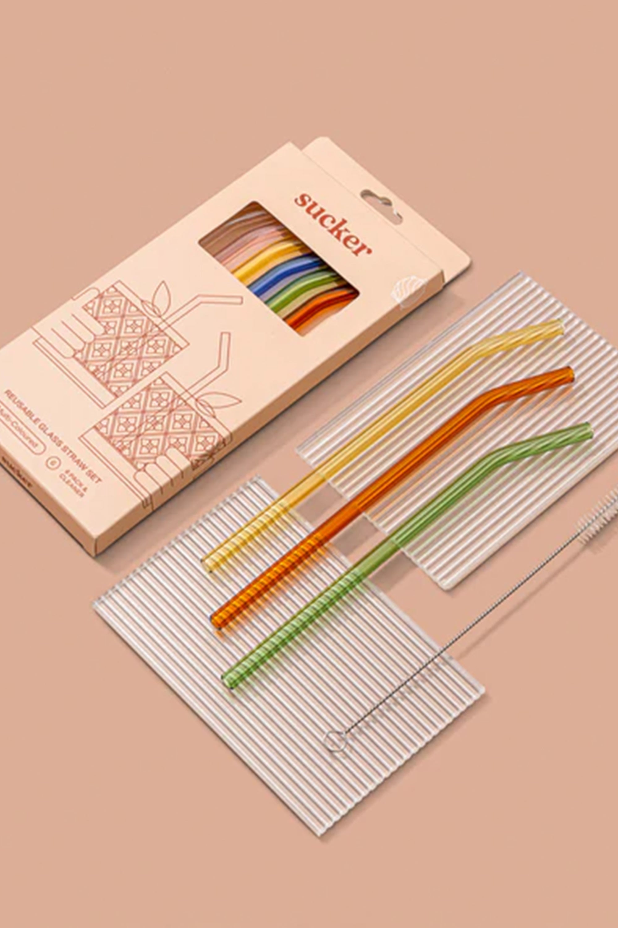 The Sucker Drinking Glass Straws by Sucker include four colorful eco-friendly straws, a cleaning brush, and come in a branded box, all shown on a beige background with two striped mats underneath.