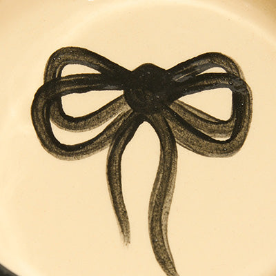 NZ-inspired black ribbon bow illustration on a cream-colored ceramic surface.